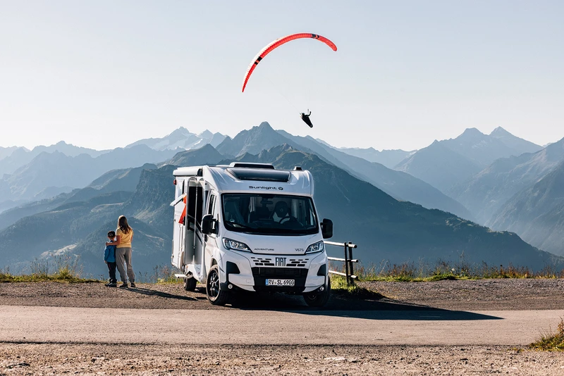 Pareja junto a una autocaravana en una carretera de montaña mirando un parapente