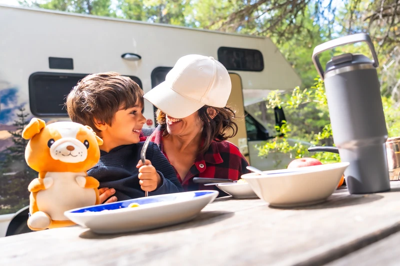 Mujer y niño merendando en una mesa de camping junto a una autocaravana