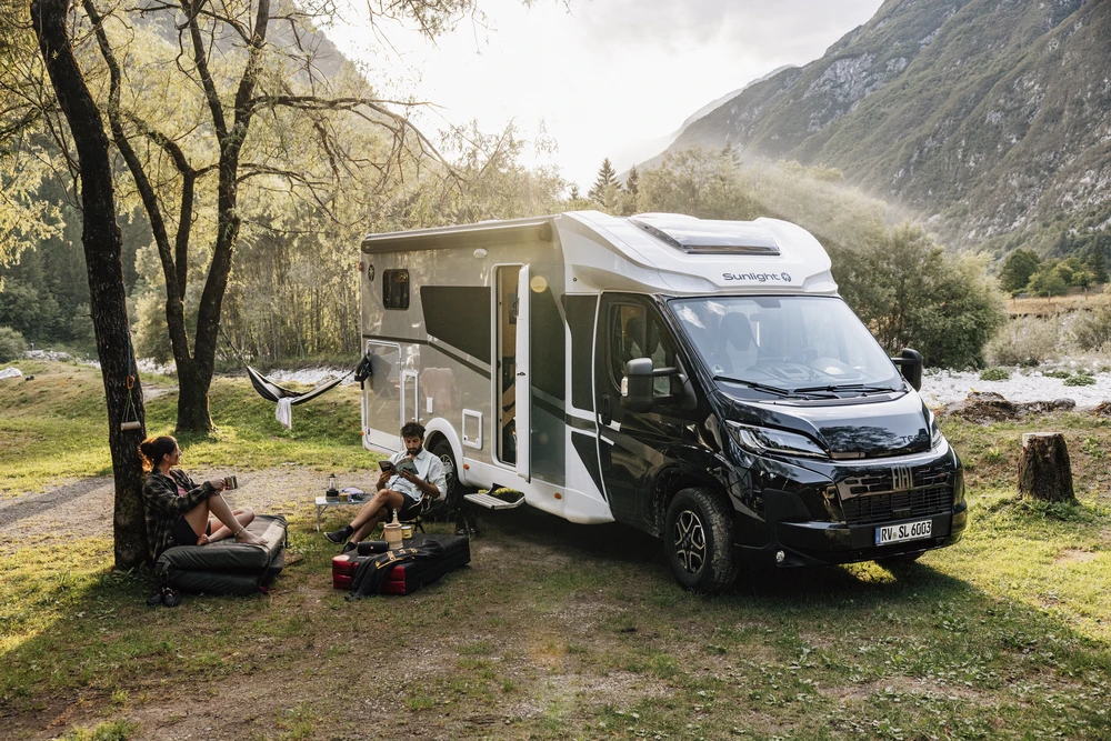 Dos viajeros descansando sobre equipo de camping junto a una autocaravana, bajo los árboles