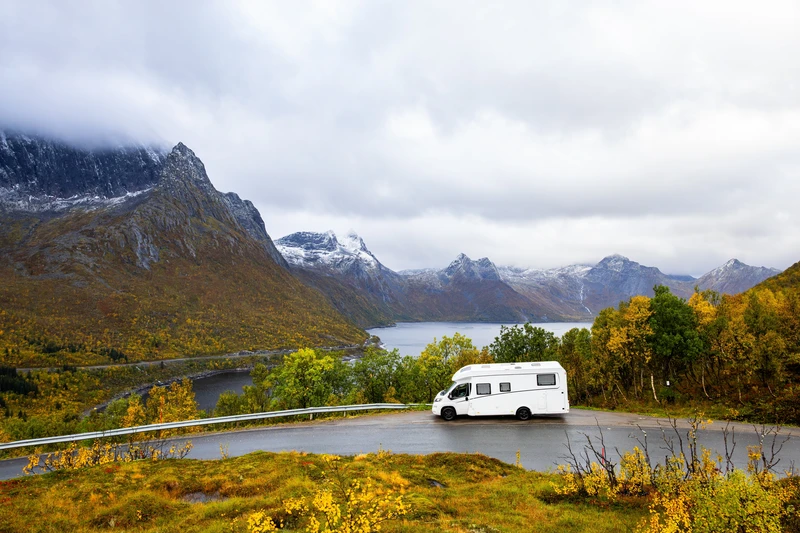 Camping-car sur une route au bord d’un lac, entre arbres d’automne et montagnes